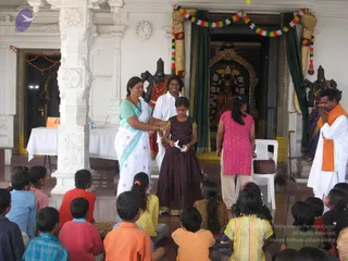 A photograph of a children's programme in a white marble temple hall. A woman in a white sari with a teal border stands at left, one hand on the shoulder of a girl in a dark maroon dress who holds a small folded paper. Behind them a man in a white shirt stands near a dark-robed deity murti in a shrine with a green curtain and garlands of multicoloured flowers. At right a man in white with an orange shawl stands beside a seated figure with a cane. A young woman in a pink kurta stands in the middle distance, and a row of seated children in bright clothes watches from the foreground. A white draped table and a small framed picture appear at left.