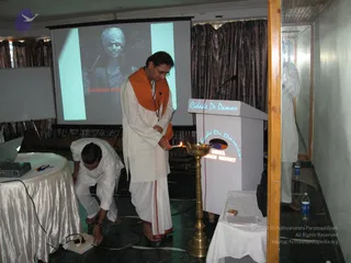 A photo taken in a conference room where a man in a white dhoti and cream kurta with an orange shawl lights a brass oil lamp on a stand beside a white podium labelled Cidade De Daman and Devka Beach Resort. Another man in white crouches at a draped table with a laptop, adjusting a power strip. On the projection screen behind them is a sepia portrait of a turbaned man captioned Architect Ana. Mineral water bottles and a box of matches sit on the small side table.