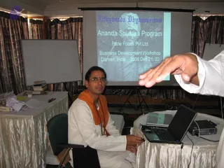 Indoor meeting room photo of a seated man in white kurta and white dhoti with an orange saffron shawl draped over his shoulders and a rudraksha mala around his neck, wearing glasses and a red tilak on his forehead, seated at a laptop on a draped table. A projector screen behind him shows a slide reading Nithyananda Dhyanapeetam, Ananda Spurana Program, Parle Foods Pvt Ltd, Business Development Workshop, Daman, India, 2006 Dec 21, 22 with a faded portrait of a turbaned figure. A blurred hand in a white sleeve gestures in the foreground, and a whiteboard, stacked books and a projector sit on nearby tables with curtains behind.