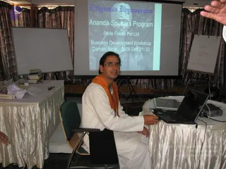 A man in a white kurta with an orange saffron shawl sits at a laptop on a draped white table in a conference room. A projector screen behind him shows a slide titled "Nithyananda Dhyanapeetam" reading "Ananda Spurana Program, Parle Foods Pvt Ltd., Business Development Workshop, Daman, India, 2006 Dec 21, 22" with a faded portrait. A whiteboard with a hand-drawn diagram and a table with papers and books are visible. The man has a tilak on his forehead and glasses, and his hand rests on the laptop touchpad. Only one person is physically present.