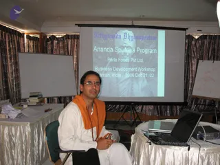 A seated man in white ritual clothing with an orange shawl and rudraksha mala speaks into a handheld microphone at a workshop. He sits in front of a projector screen showing a slide titled "Nithyananda Dhyanapeetam" and "Ananda Spurana Program," with a sepia portrait of a turbaned figure. Behind him are brown patterned curtains, a whiteboard on the left, and a silver-draped table with books, papers and a laptop on the right, plus a flipchart with a sketch on the far right.