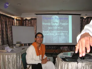 A seated man in white kurta and white dhoti with an orange-saffron shawl draped over his shoulders and a rudraksha mala around his neck holds a microphone while sitting in a meeting room. A projection screen behind him shows a slide with the title 'Nithyananda Dhyanapeetam' and 'Ananda Spurana Program', with a sepia photograph of a turbaned man in white robes. A laptop, projector, books and papers sit on a draped table to the left, and a blurred hand reaches into the right foreground. Curtains and a whiteboard line the room.