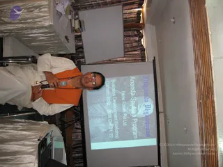 A photograph taken in a hall with the image rotated sideways. A man stands at left holding a microphone in his right hand, wearing a white kurta, an orange shawl over his shoulders and a rudraksha mala around his neck, with glasses on. Behind him a projector screen shows a slide reading Ananda Sputana Program with a photo of a man and text about a business development workshop in Daman, India, Dec 21-22, 2006. A white-covered table, a laptop and a curtained window are in the room.