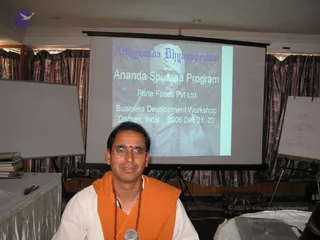 A man seated in front of a projector screen in a hotel-style meeting room, wearing an orange shawl over white clothes and a rudraksha mala, with a red bindi on his forehead, glasses, and a microphone in front of him. The projected slide reads Nithyananda Dhyanapeetam, Ananda Spurana Program, Parle Foods Pvt Ltd., Business Development Workshop, Daman, India, 2006 Dec 21, 22, with a black-and-white portrait of a turbaned figure beside the text. Curtains, a whiteboard with a hand-drawn sketch, and a table with books are visible. The seated man is not clearly identifiable from appearance alone.