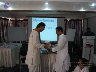 Indoor event photo in a meeting hall with curtains and a projector screen reading "Nithyananda Dhyanapeetam" and "Parle". Two men stand at a central table exchanging a stack of books; the man on the left wears a white kurta, an orange shawl and glasses, and the man on the right wears a white kurta. A white lectern labelled "Cidvilasa De Daman" and "Devika Beach Resort" stands at right, with an oil lamp on it. A man in a dark shirt stands behind the lectern, and a seated person in white is partly visible at lower right. Whiteboards and a draped table with papers and a laptop are present.