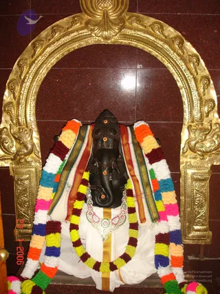 A dark stone Ganesha murti stands in a gold-framed arched niche on a polished maroon granite wall. The elephant-headed deity wears a white dhoti with a gold border, a multicoloured striped sash, and a silver-and-jewelled necklace. Multicoloured flower garlands in orange, white, yellow, maroon and blue hang from its shoulders, and red tilak marks decorate its forehead and trunk. The ornate gold arch is embossed with floral and peacock-like motifs. No people are visible.
