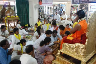 A photograph of a padapuja-style ceremony in an indoor hall lined with bookshelves of books and DVDs. A seated figure in an orange saffron robe with a flower garland sits on an ornate gold throne at right, hands folded in front, with a striped red and orange cloth draped over the throne's side. Several people in white clothing and yellow shawls sit on the floor in front of the throne, while a woman in a lavender sari and others in white kneel forward with flowers and brass plates of fruit and offerings. Overhead, a wooden canopy stands at left, decorated with yellow and orange marigold garlands, and a banner reading Books, DVDs, Audio etc. are available hangs above. Several seated devotees in white clothing press their palms together in devotion.