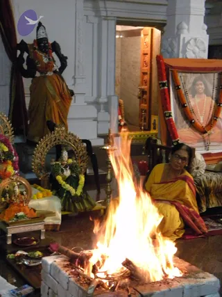 A woman in a yellow saree with a red border sits beside a homa fire pit lined with white bricks, flames rising high in the centre. Behind her, a framed portrait of a smiling man with long hair in orange robes and a rudraksha mala is garlanded with orange and red flowers under an orange canopy; the portrait is a depiction, not a physical person. A dark stone deity murti stands in a sari, a brass aarti lamp and oil lamps stand near the fire, and brass plates hold flowers and offerings on a small table. The woman is the only physical person visible.