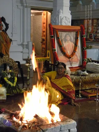 A woman in a yellow sari with a red-and-maroon border sits on the floor beside a stone-lined fire pit where a tall flame rises from burning logs. She wears glasses and a necklace and reaches toward the fire. Behind her a framed portrait of a long-haired figure in saffron robes, with white beads and rudraksha malas, is draped with a saffron cloth and garlanded with orange and red flower garlands. The portrait is labelled in small printed text. A tiger-striped cushion rests nearby, with brass oil lamps and a brass kalasha-like vessel. On the left, a deity murti in a gold arch is decorated with a green and yellow garland. The setting looks like a homa in a temple hall.