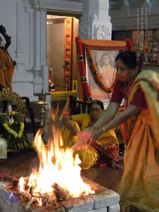 A woman in a rust-orange sari with a dark red blouse leans over a stone-lined fire pit, extending her hands toward a bright open flame in an indoor temple-like hall. Behind her, a seated woman in a yellow sari and glasses watches the fire. In the background, a portrait of a smiling man with long hair, wearing an orange robe and white flower garland, is framed by a canopy with red and orange marigold garlands and rudraksha beads. A brass oil lamp stands nearby, and a deity murti adorned with green and yellow garlands sits at left beside white pillars. The scene appears to be a fire ritual being performed at a temple shrine.