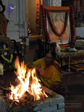 A woman in a yellow saree with dark red border kneels beside a brick-lined fire pit, feeding wood into a bright blazing homa fire in a temple hall. Behind her, a framed portrait labelled "Paramahamsa Nithyananda" is draped with orange cloth and garlanded with red and orange flower garlands and rudraksha malas. The portrait shows a smiling figure in orange robes with white beaded necklaces, seated in meditation. Nearby stand brass oil lamps, a deity murti with yellow flower garland, and a tiger-striped cushioned seat.