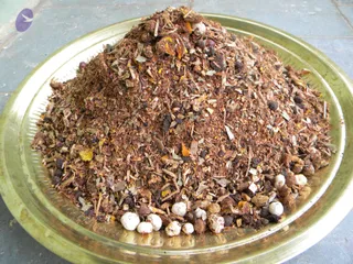 A close-up photo of a heaped mound of dried, crushed herbal material and ground powder piled on a round brass plate with a raised rim. The mix includes dark brown powder, dried leaf and bark fragments, small dark seeds and pieces, orange and yellow flecks, and several whitish round seeds clustered at the lower edge. The plate rests on a grey tiled surface. No people are visible.