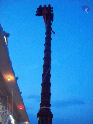 Low-angle photo at dusk of a tall dark metal flagstaff (dhwajastambha) rising against a deep blue evening sky. Its top bears an ornate finial with a small red pennant fluttering from the upper section, and a thin cable runs along the pole. A tiled temple roof edge with lit lamps and ornamental carvings appears at the left, with a warm light glowing beneath the eave. No people are visible.