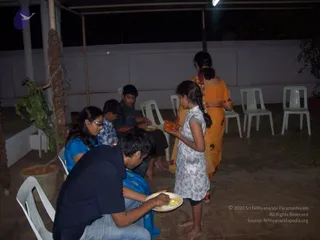 A night-time photo on an open-air concrete terrace with white plastic chairs and a rope-wrapped pillar at left. A woman in a bright orange sari with a long braid stands at right, and a girl in a white sleeveless dress with a braid holds a small plate and a leaf or packet. Four other people are seated or crouching at left: a woman in a blue top and glasses, a young boy in a blue shirt, an older boy in dark clothes, and a man in a navy shirt eating from a plate of food. A dim overhead light and a plant in a pot complete the scene.