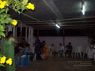 A night-time photograph of a covered open-sided hall with a corrugated metal roof and two fluorescent tube lights. Several people sit on white plastic chairs eating from plates, while others stand nearby. A woman in a green sari stands at left beside yellow flowering branches, and a woman in blue stands in the centre. A man in a white shirt sits at the far right holding a plate. The setting looks like a simple communal meal, with no visible deity, throne or ritual objects, and no visible a person candidate.
