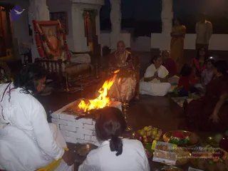 A photograph of a homa at dusk in a temple hall with white pillars. A brick fire pit burns brightly in the centre. A woman in a gold-bordered sari sits on a wooden chair behind the fire. Nearby, a woman in white with folded hands sits on the floor, and a man in a red shirt sits beside her. Other devotees sit or stand around the fire, including a woman in a dark red sari on the right and two girls in red and pink. On the left, a woman in white with her back to the camera faces the fire. A decorated empty seat with red and white garlands and a framed portrait stands at left, beside a brass lamp. Brass plates of fruit, flowers and sweets sit in the foreground.