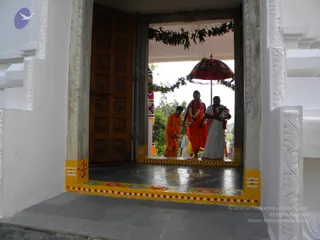 A photograph taken through a white temple doorway with a carved wooden door on the left. Three people walk across a dark tiled floor toward the camera: a figure in orange robes bending slightly at left, a central figure in a deep orange robe with a white flower garland, and a figure in white dhoti and shawl with a red border carrying items. A red and gold umbrella hangs overhead, and green leaf festoons decorate the doorway lintel. Yellow-painted door frames bear an om symbol and red and white dotted borders. Greenery is visible beyond the entrance.