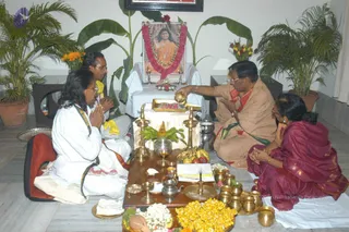 Indoor puja before a framed portrait of a saffron-robed figure with long hair and rudraksha beads, garlanded with red and white flowers and flanked by two lit oil lamps on a white-draped table. Four people sit on the floor: a bearded man in white kurta with folded hands at left, a man in white with a yellow shawl behind him, an older man in beige and orange robes pouring water from a brass vessel, and a woman in a maroon sari with folded hands at right. Before them stand brass lamps, a kalasha-style vessel, fruit, flower garlands, a coconut, and a heap of yellow grain. Potted palms and a banana plant fill the room.