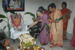 Indoor puja before a framed portrait of a man in saffron robes with a long garland of red and pink flowers, placed on a table draped in white cloth. The portrait is labelled "Paramahamsa Nithyananda" on its caption. An elderly woman in a purple sari with green border reaches toward the offerings while a man in a pale peach dhoti and shawl with a green border, holding a small book, stands beside her. A woman in a red floral sari stands at right, and a woman at lower left presses her palms together in prayer. A brass oil lamp, a flower-decorated plate, bananas and fruit sit on a tray in front. A framed painting of a deity hangs on the wall above, with a large green plant beside it.