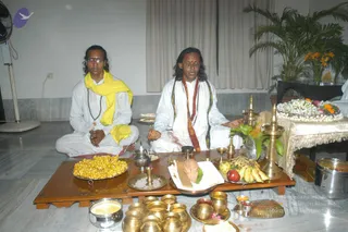 Two men sit cross-legged on a marble floor before a low wooden puja table, performing a ritual with eyes partly closed. The man on the left wears white clothing, a yellow shawl, glasses and a rudraksha mala. The man on the right, in white with a red-and-gold shawl and rudraksha mala, has a vibhuti or kumkum mark on his forehead and holds his hands in a mudra over the offerings. The table holds bananas, apples, a yellow sweet in a platter, a brass kalasha, brass lamps, a bowl of grains, a coconut-like offering on a leaf, and small brass vessels. A garland of white and orange flowers rests on a side table, and a palm plant stands behind. A ceiling fan is at left, and a steel pot sits at lower right.