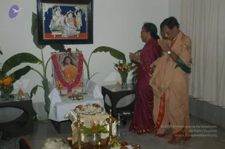 A domestic puja room with a framed tanjore-style painting of a deity trio on the wall above. Beneath it, a framed portrait labelled "Paramahamsa Nithyananda" sits on a white-draped stand, garlanded with a pink and white flower garland and framed by oil lamps. The portrait shows a figure in orange robes with a long-haired, wide-set look and a beaded necklace. In front stands a brass tray with flower garlands, fruit, a kalasha-style vessel and brass oil lamps. Two standing people face the altar with palms pressed together in namaste: a woman in a maroon saree with a plait, and a man in a peach-coloured dhoti and shawl with a green border and a wristwatch. Potted plants and vases of orange and red flowers frame the scene.