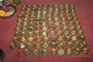 A photograph of a homa preparation laid out on a red cloth floor covering. Rows of small brass bowls, roughly a dozen across and about ten rows deep, each hold a mix of dried herbs, bark pieces, seeds, grains and small white items, arranged in a neat grid. In the upper left, a brass bowl holds marigold and yellow flowers, red petals and green leaves, beside a coconut half and leaves on a tray. No people are visible in the frame.