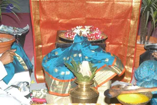 A ritual setup photographed indoors. A large deep-orange and gold brocade cloth hangs behind a low dark wooden stand holding a brass plate heaped with red and white flower garlands and a small brass lamp. In front, a teal-blue silk cloth with gold square motifs and orange-and-gold border is draped over a mound, possibly a sacred object covered for worship. A brass kalasha/pot with mango leaves and a white tuft sits in the foreground, with a wooden spoon in a bowl of yellow liquid at right. At left, a partially visible person in white clothing holds the teal cloth edge, and at right another person in a teal patterned garment is partly visible. Potted plants stand at both sides.