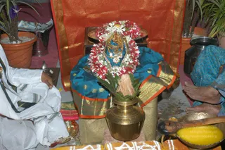 A photograph of a ritual worship scene in front of an orange and gold-bordered cloth. At centre, a deity murti or image draped in a teal silk cloth with gold zari borders is adorned with a large garland of red and white flowers and gold-toned ornaments. A brass kalasha or pot with green leaves stands in front, with a brass plate of offerings behind. On the left, a person in white ritual clothes sits with a hand near a brass lamp. On the right, an elderly woman in a blue sari sits barefoot near brass vessels and a yellow-filled bowl. Potted plants and a brass vessel frame the scene, and colourful kolam or powder designs lie on the floor below.