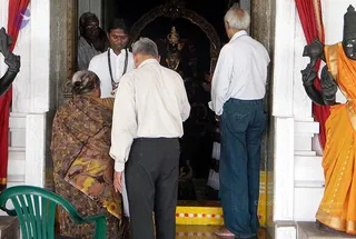Devotees enter the doorway of a temple shrine with a dark deity murti visible inside, framed by a gold arch. In the foreground, an older man in a white shirt and dark trousers bends forward, and a woman in a brown and gold patterned shawl stands at left. A younger man in white with a dark beaded mala stands in the doorway, and an older man in a white shirt and jeans stands at right. A red curtain hangs at the sides, and a large stone dvarapala figure in an orange-gold garment stands on the right. A green plastic chair sits at lower left. The people appear to be taking darshan at the shrine threshold.