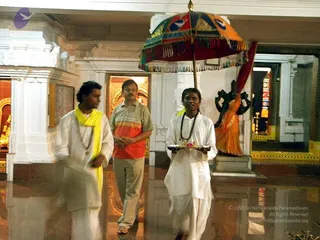 Inside a white marble temple hall with a polished reflective floor, a young man in white dhoti and shirt with a rudraksha mala carries a silver tray with offerings or lamps, while a man in a yellow-and-orange striped shirt and a second man in white with a yellow shawl walk alongside. Behind them, a large red, orange and green ceremonial umbrella with fringe is held aloft, and a dark deity murti in an orange-draped niche stands near gilded doorways.