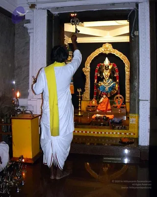 A person seen from behind performs aarti with a lit lamp held high in the right hand before a shrine set in a golden arch. The person wears white dhoti-style clothing with a yellow shawl over one shoulder. Inside the shrine stands a decorated deity murti dressed in blue and gold with red garlands, with a small orange-clad figure seated before it on a dais. Brass oil lamps with lit flames stand on the left and in front of the shrine, and a plate of offerings sits at the right edge. The setting is a temple interior with white marble pillars and a glossy dark floor.