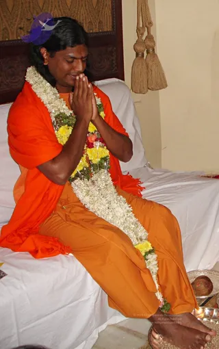 A man seated on a white-draped bed with a dark wooden headboard, hands pressed together in anjali mudra with eyes lowered. He wears an orange-saffron shawl over orange robes and a long garland of white jasmine and yellow marigolds with red flowers draped from his neck to his lap. A small red tilak is visible on his forehead. Behind him hang tasselled cords, and at his feet stand a silver plate with a coconut and a brass tray. a person by appearance: long swept-back hair, saffron clothing and ceremonial garland, seated as the one being honoured.