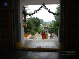 Photograph taken from inside an ornate wooden temple doorway looking out at a broad stone staircase ascending between white and red railings toward green trees and open sky. A green leafy garland arches over the entrance, and a painted pillar wrapped in a colourful patterned cloth with blue bird motifs stands at the left. Several people in saffron and orange robes, a woman in red with a garland, and a person in white climb the steps; a person in white holds a tray. A multicoloured striped umbrella is held over the steps. Decorative white and yellow rangoli designs and a red floral kolam mark the threshold floor, and a faint Om symbol appears on the yellow door frame.