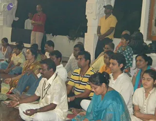 Indoor photo of devotees seated on mats in a white-pillared hall at night, eyes closed in meditation or chanting. In the foreground a man in white kurta with rudraksha mala and a woman in blue salwar kameez sit with hands in mudra. Others in yellow and striped shirts, white kurtas and saris sit nearby, while two men stand at the back, one in red shirt and one in yellow shirt with cap. White carved pillars frame the hall and a dark backdrop rises behind. No clear depiction of a specific guru is visible.