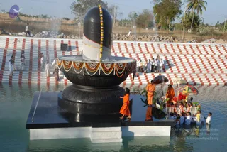 Outdoor photo of a large black Shiva lingam with white stripe and marigold garlands on a black yoni pedestal, standing on a platform in a water tank. Water sprays from the base. Behind, white and red striped steps hold onlookers in white and light clothes. In the foreground, a figure in orange robes stands near the lingam while another in orange with a long wrap and a shaved head stands on the platform, with a boat decorated with garlands and orange-and-colourful umbrellas carrying people in orange and white. Several people in white clothes stand in the shallow water near the boat. Palm trees and dry scrub surround the site.