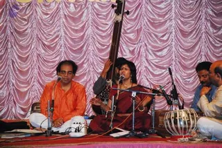 A stage performance before a pink draped backdrop with swagged fabric. A man in an orange-saffron kurta sits cross-legged on the left, with a microphone in front of him. Beside him, a woman in a deep maroon outfit with gold jewelry sits playing a long-necked tanpura or veena-style instrument upright, with a microphone stand near her. Two men sit at the right, one in a light blue shirt and another in white, with a tabla drum set on the floor in the foreground. Carpeted stage floor with red and pink tones.