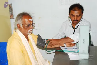A medical check-up in a plain room with a white cinder-block wall. An older man with white hair, white moustache, glasses and a vibhuti mark on his forehead sits on a blue plastic chair, wearing a yellow kurta and a white shawl. A young man in a white striped shirt with a stethoscope around his neck wraps a dark grey cuff on the older man's upper arm and checks his blood pressure on a Life-Line monitor placed on a metal table with papers and a pen. The older man is not identifiable as a person from visible cues here, so no candidate is asserted.