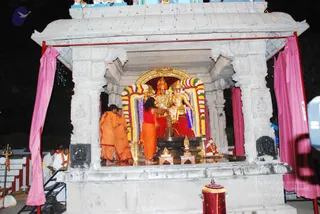 A night photograph of an outdoor abhishekam at a white marble temple-style shrine decorated with pink cloth hangings and string lights. At the centre, two deity murtis sit on an ornate gold-framed pedestal, adorned with garlands of marigold and mixed flowers. In front, a figure in an orange robe stands at a central stone lingam-style vessel pouring liquid, while two men in orange and saffron clothing stand nearby. Another man in white garments and orange sash stands at the lower left beside a brass lamp stand and a trident-like brass object. A red and gold cylindrical item sits at the front of the platform. Lights are strung along the top of the structure.