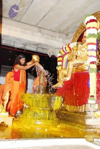 A woman in a red blouse and orange saffron sari stands at left, pouring water from a brass kalasha over a golden deity murti seated on a throne. The murti wears a red garment and is draped with garlands of white, red and yellow flowers. Water splashes down onto a yellow-tinted pedestal. A second person in orange robes stands behind her at left. The setting appears to be a temple interior with a concrete ceiling. This is an abhishekam ritual.