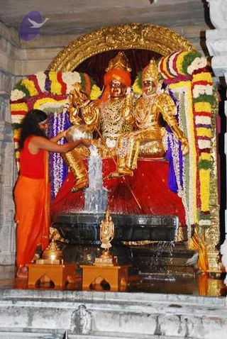 A photograph inside a temple shows a golden deity murti of a seated couple, one figure in an orange-red turban and the other with a tall gold crown, both draped in gold ornaments and sitting on a red cushioned seat above a black stone pedestal. Garlands of yellow and orange flowers with green leaves frame the shrine arch. A person in an orange-red top and orange dhoti stands at left, pouring liquid from a metal vessel over the murti, with water flowing down onto the black base. Brass oil lamps and a small brass vessel sit on the floor in front. This is an abhishekam offering.