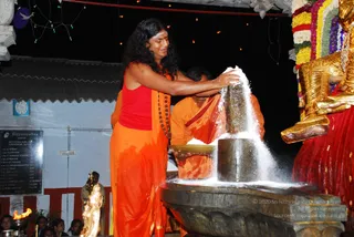 Night ritual photograph at a temple shrine. A long-haired man in an orange dhoti and red sleeveless top, with a rudraksha mala around his neck, pours white granular material from a bowl over a dark stone Shivalinga set in a basin. Behind him, a second man in orange robes holds a brass plate. To the right, a large golden deity murti wearing a red garment and flower garlands stands beside the shrine. Strings of small lights hang overhead, and a brass lamp with a flame sits at the lower left among seated devotees. A plaque on the wall lists devotee names in the Dhyanapeetam shrine. This appears to be an abhishekam, the ritual bathing of the Shivalinga.