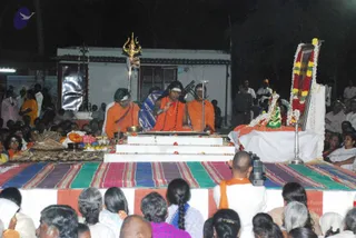 Night-time outdoor ritual on a raised white platform with a striped multicolour mat. Three central figures in saffron-orange robes sit on the dais; the central figure wears a rudraksha mala and a white tilak, with ornate gold necklaces, and gestures with his hand while speaking into a microphone. Beside him stands a golden trident on a pole, and in front are brass vessels, a flat bowl, and offerings of flowers and leaves, consistent with an abhishekam-style puja. To the right, a framed picture decorated with a garland of white and orange flowers and green leaves sits on a draped table. Several dozen seated devotees and a cameraman in orange fill the foreground, with onlookers standing behind a white building in the background.
