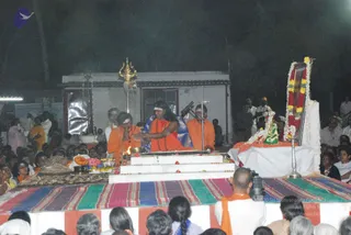 Night outdoor ritual on a raised white platform draped with striped cloth. A central figure with long dark hair, a white headband and a blue shawl over orange clothing extends his hands over a small fire pit; two other orange-robed priests stand nearby, one holding his hands together. Brass oil lamps, flower offerings, fruit and a brass trident stand behind on the platform. To the right a deity murti or framed image is decorated with marigold garlands and a green cloth. Seated devotees fill the foreground and background, and a cameraman in orange clothing films from the front.