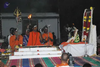 Night ritual outdoors on a striped woven mat before a white-draped low platform. A central figure with long dark hair, a white forehead band, a red-orange robe, a rudraksha mala and a gold chain holds a lit torch upward in one hand. Two attendants in orange robes with white forehead marks stand beside him; the one on the left holds his hands together in prayer, the one on the right holds a ritual implement. A golden trident stands on a pole at left, a garlanded framed portrait of a deity with marigold and yellow flower garlands sits at right, and fruit and flower offerings lie on a tray in the foreground. Several standing onlookers in white and dark clothing watch in the background, and a camera operator's head is visible at the bottom.