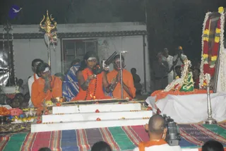 Night outdoor ritual photo. At the centre, a bare-chested figure in saffron-orange cloth with long dark hair, rudraksha mala beads and a white-marked forehead holds a ceremonial implement over a fire on a white stepped platform. Beside him, a man in orange robes with a white-smeared forehead presses his palms together, and another man in orange stands behind with hands folded. A young man in orange kneels to the left with hands joined. A large flower-garlanded framed portrait stands at the right on a white-draped altar, with a green cloth, yellow flowers and a brass lamp; a trident stands behind the platform. A cameraman in orange sits at the bottom. Crowd members stand in the background.