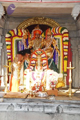 A photograph inside a stone temple shrine showing a golden deity pair seated under an arched gilded frame, draped with garlands in yellow, red, white, purple and green. The central figure wears an orange turban and rich necklaces, with a trident-like staff at his side, and the consort figure wears a gold crown. In front, a large golden lingam is decorated with flower garlands and sits on a brightly lit platform. A woman in a gold and leopard-print dress stands at left with palms pressed together in prayer. Brass lamps stand on either side, and a silver offering vessel and small brass bowls sit on the platform. No a person candidate is identifiable.