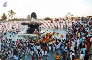 A large crowd stands in shallow water around a tall black Shivalingam set on a raised black pedestal within a rectangular tank. Many people wear white clothing, and others wear bright orange, yellow and pink garments. A decorated palanquin or chariot with red, orange and gold ornaments and umbrella-like canopies is carried through the water at the centre. Behind the tank stand red-and-white striped walls, palm trees and more spectators along the edge.