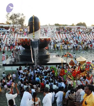 Large outdoor photo of a giant black Shiva lingam with a yoni-shaped base at a temple pool, water flowing down its shaft. Devotees in white clothes fill the shallow water around the lingam and the crowd lines tiered red-and-white seating on the steps behind. Adjacent to the water, decorated deity idols on a colourful chariot with multicoloured canopies and umbrellas are surrounded by people. A man in a yellow shirt in the lower right raises a hand, and a priest in white stands at the left edge beside the lingam. Crowd of many devotees visible.