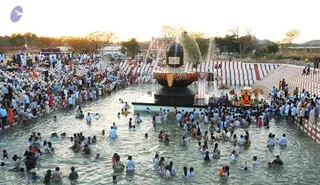 Wide outdoor photograph of a large crowd bathing in a shallow stone-edged pool at dusk. In the centre stands a huge black stone Shivalinga with an orange and gold decorated base, with water jets spraying around it. Many people wearing white and light clothing stand waist-deep in the water, some pouring water and folding hands in devotion. Along the pool edges, red and white striped tent-like structures and rows of people in white line the sides, with trees and a pale sky behind.