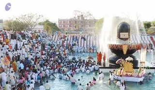 A large outdoor ritual at a temple-style complex, with a big crowd gathered around a shallow pool. A large black stone Shiva lingam with a golden base and decorated gold panels stands in the water at the right, beside a golden kalasha-style structure, with fountain sprays rising behind it. Many people in white clothes wade in the pool, some submerged to the waist, while others stand on the surrounding paved edge. Among the crowd, a few saffron-robed figures in orange stand near the lingam, and several people carry flags and ornaments. Striped red-and-white canopies and a brick building appear in the background under a pale sky.